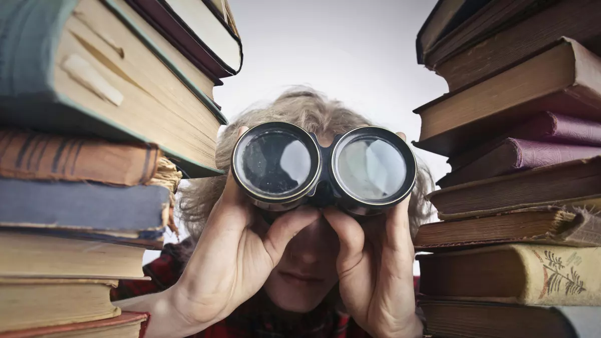 A person with blonde hair looks through binoculars, surrounded by a stack of books.