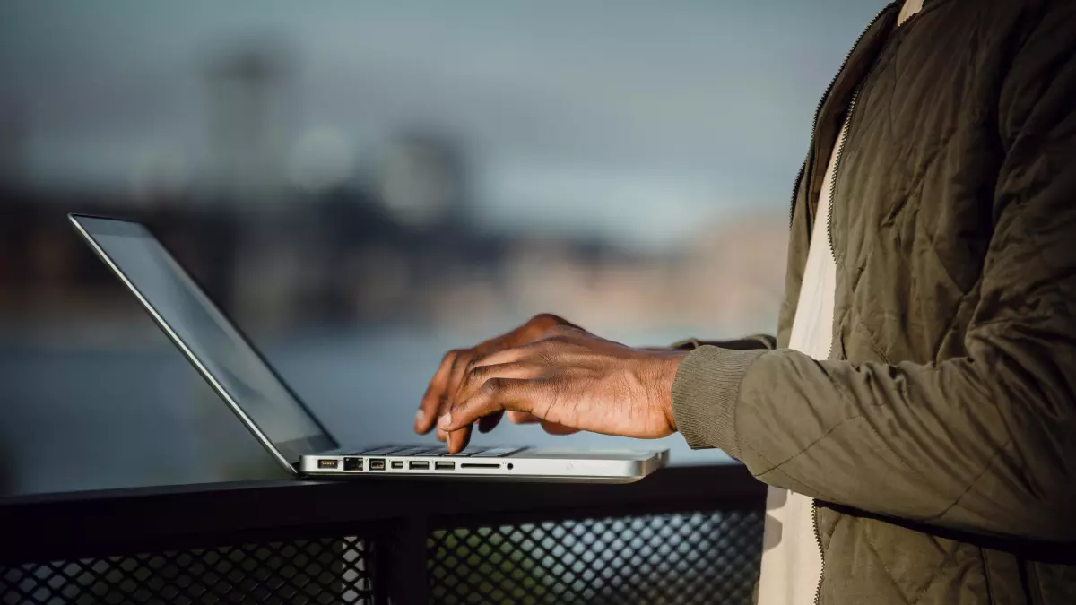 A person working on a laptop with their back turned to the camera. They are standing on a balcony, and the background is blurred.