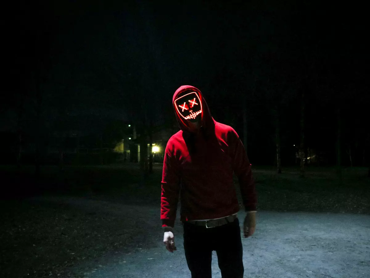 A man is wearing a red hoodie and a glowing mask in a dark environment.