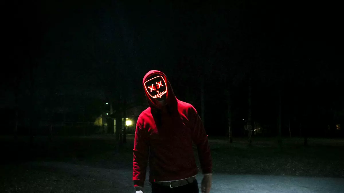 A man is wearing a red hoodie and a glowing mask in a dark environment.