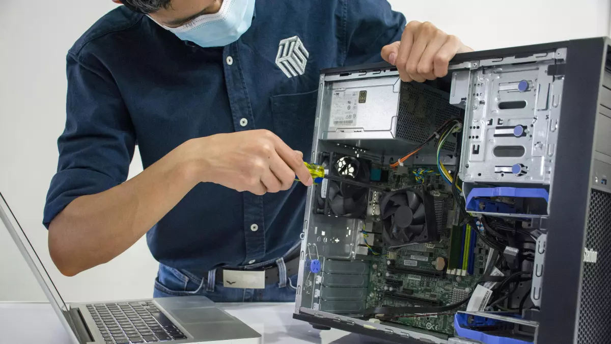 A person in a blue shirt and face mask is working on a desktop computer, connecting a cable. There is a laptop in front of him.