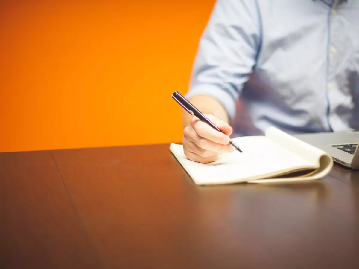 A person is sitting at a desk, writing in a notebook with a pen. A laptop computer is on the table next to them. The background is a bright orange wall. 