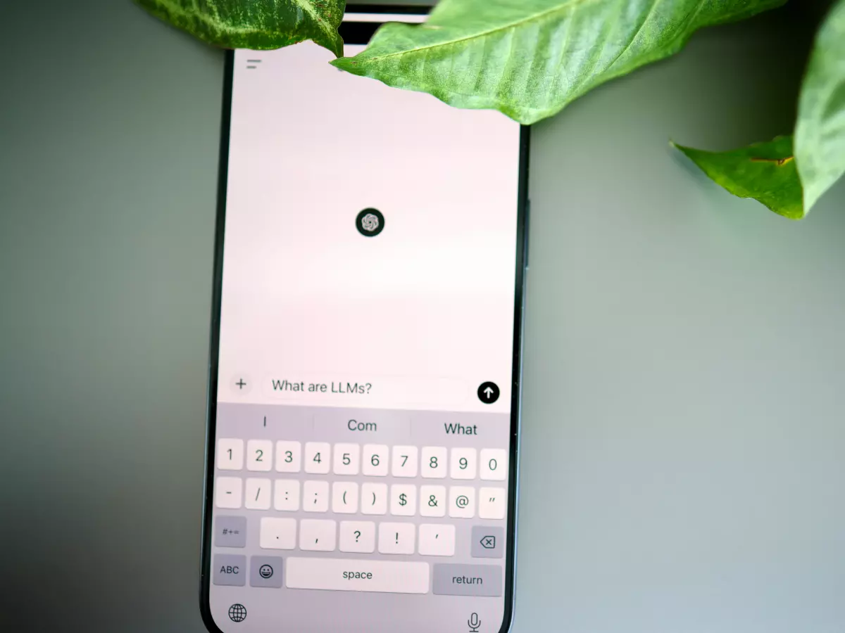 A smartphone displaying a text input screen with a keyboard; a leafy plant is partially obscuring the top of the screen.