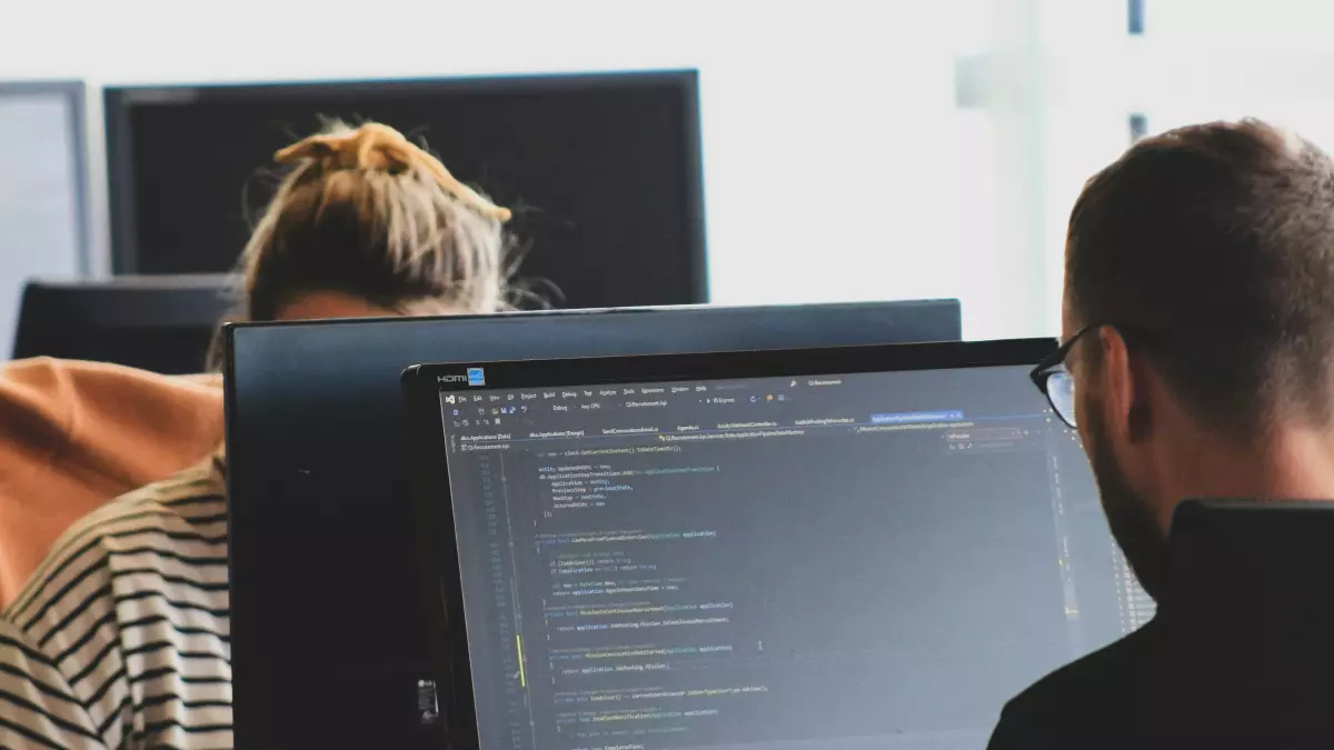 Two people are working on their computers, focused on lines of code. The background is a bright office space.