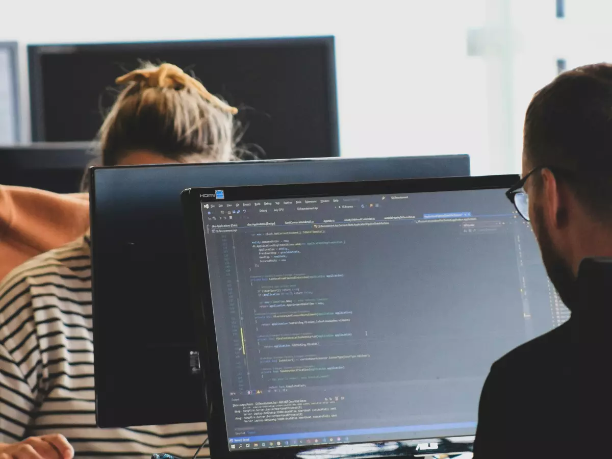 Two people are working on their computers, focused on lines of code. The background is a bright office space.