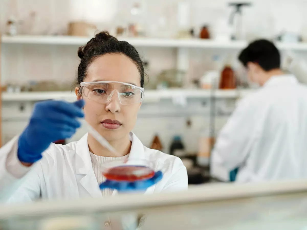 A female scientist in a lab coat and protective eyewear carefully pipettes a liquid sample into a petri dish.