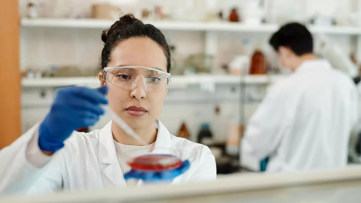 A female scientist in a lab coat and protective eyewear carefully pipettes a liquid sample into a petri dish.