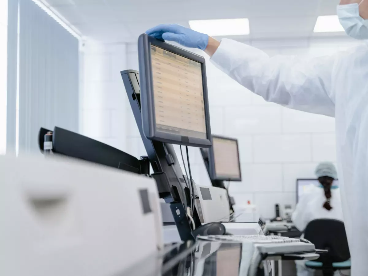 A scientist in a lab coat adjusts a computer monitor displaying data; multiple monitors and lab equipment in the background.