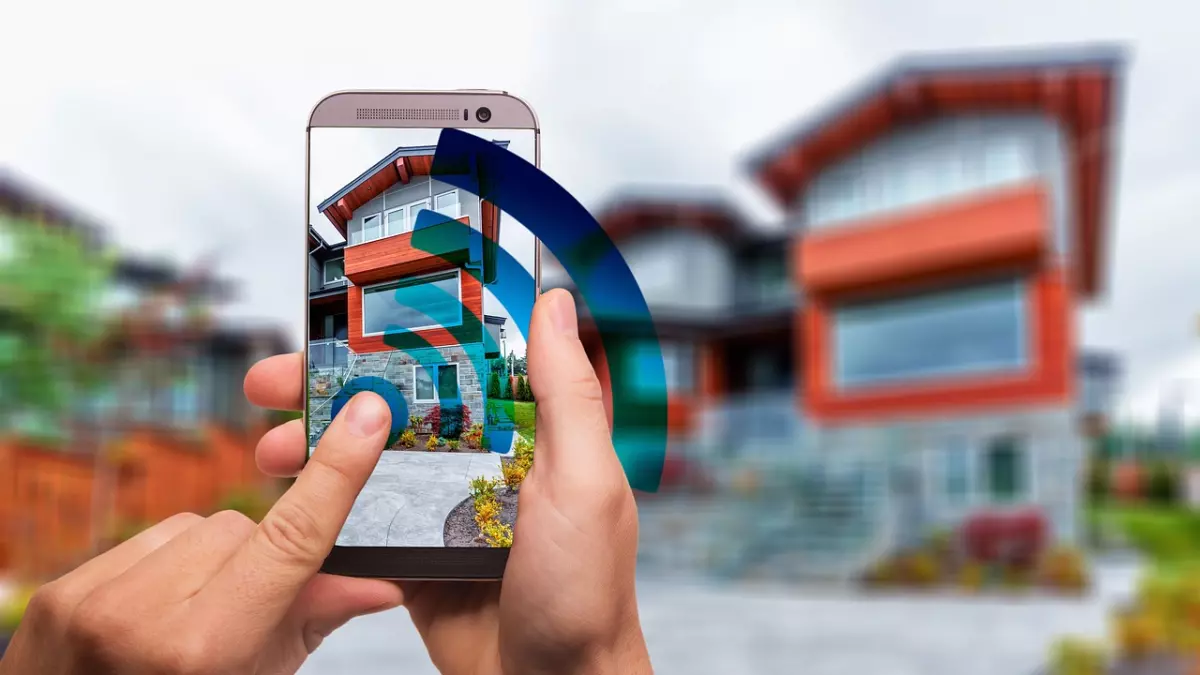 A person is holding a smartphone in their hands. The phone is displaying a house with a blue wifi signal around it. The house is in the background, out of focus.