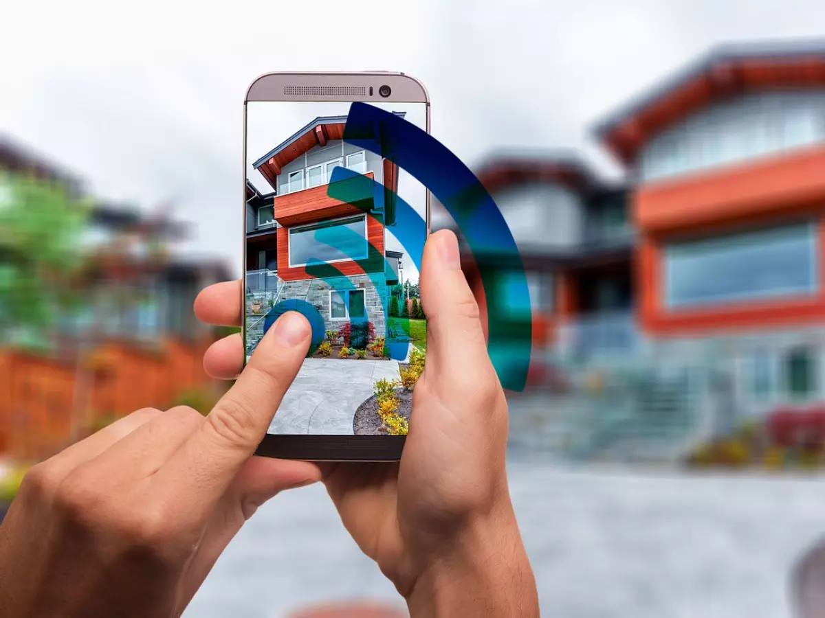 A person is holding a smartphone in their hands. The phone is displaying a house with a blue wifi signal around it. The house is in the background, out of focus.