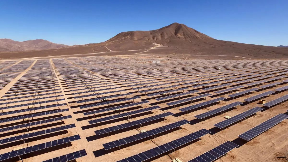 An expansive desert solar farm stretches across a vast landscape under a clear blue sky, with mountains in the distance.