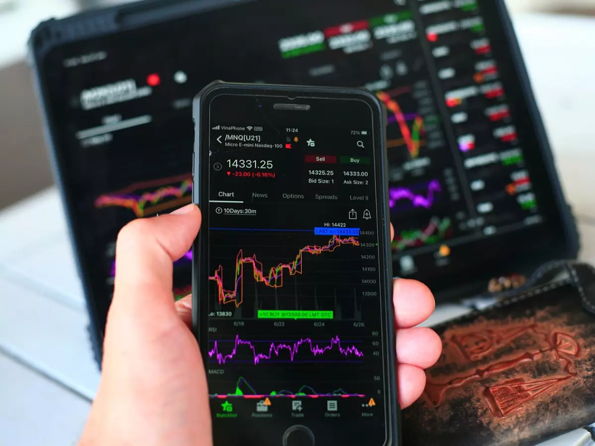 A close-up shot of a hand holding a smartphone displaying stock market data, with a laptop screen in the background showing similar data.