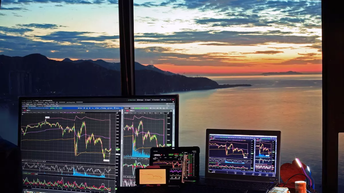 A desk with multiple devices displaying stock market charts. The scene is set in a room with a large window offering a panoramic view of a cityscape at sunset. The desk is also equipped with a keyboard, mouse, and other accessories, highlighting the digital nature of the subject matter.