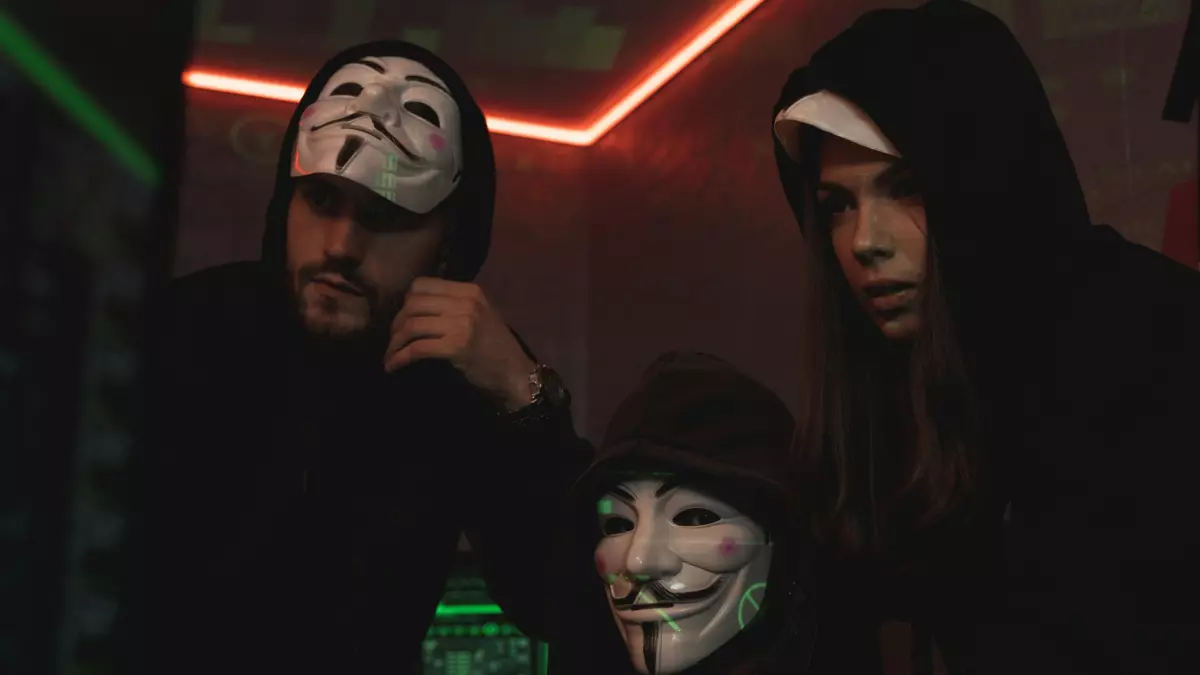 Three people wearing black hoodies and masks are looking at a computer screen, the image is shot from a low angle.