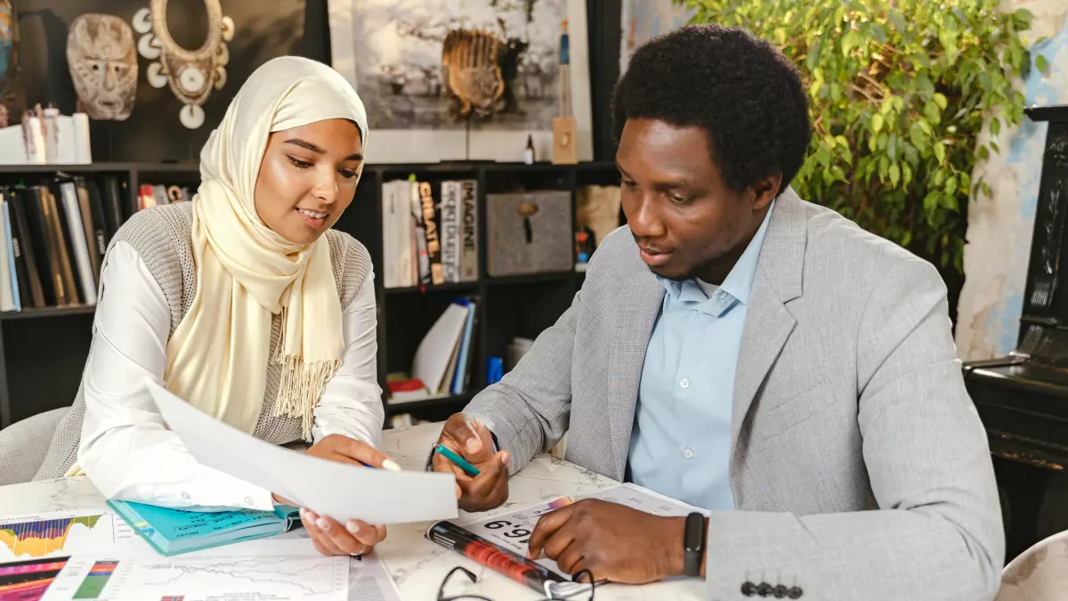 Two people, a woman in a headscarf and a man, are sitting at a table and looking at papers, both of them appear engaged in a project.