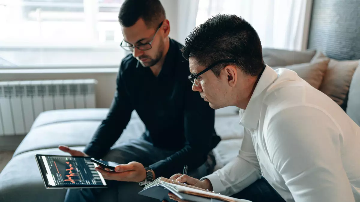 Two men sitting on a bed, looking at a tablet and a phone, discussing something