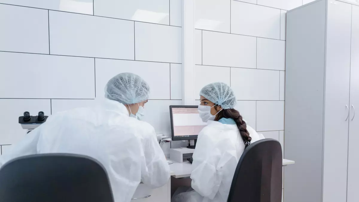 Two people in lab coats are sitting at a computer, looking at a screen with graphs.