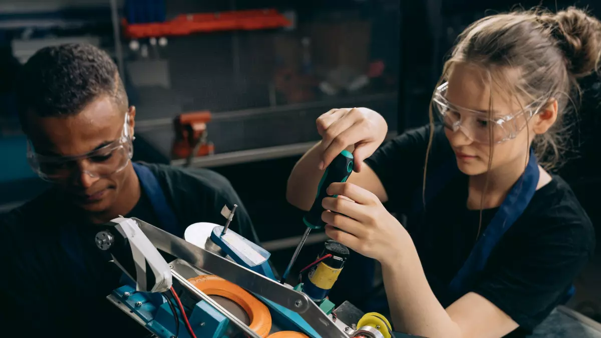 Two people, a man and a woman, are working on a robotic device. They are wearing safety glasses and focused on their task. The setting appears to be a workshop.