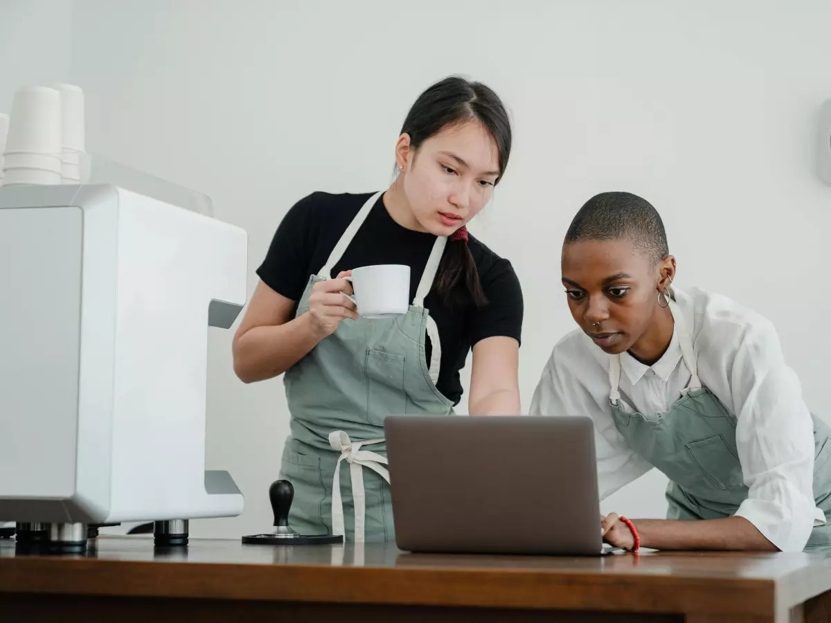 Two people, one wearing a black t-shirt and a green apron, and the other wearing a white shirt, are looking at a laptop together.