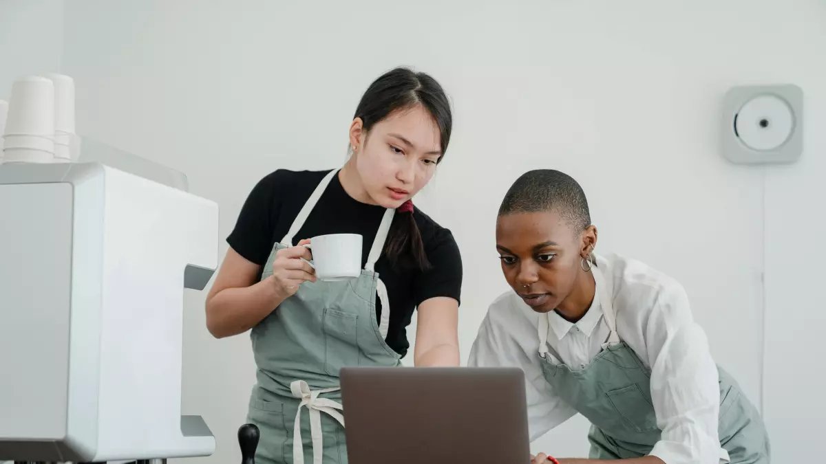 Two people, one wearing a black t-shirt and a green apron, and the other wearing a white shirt, are looking at a laptop together.