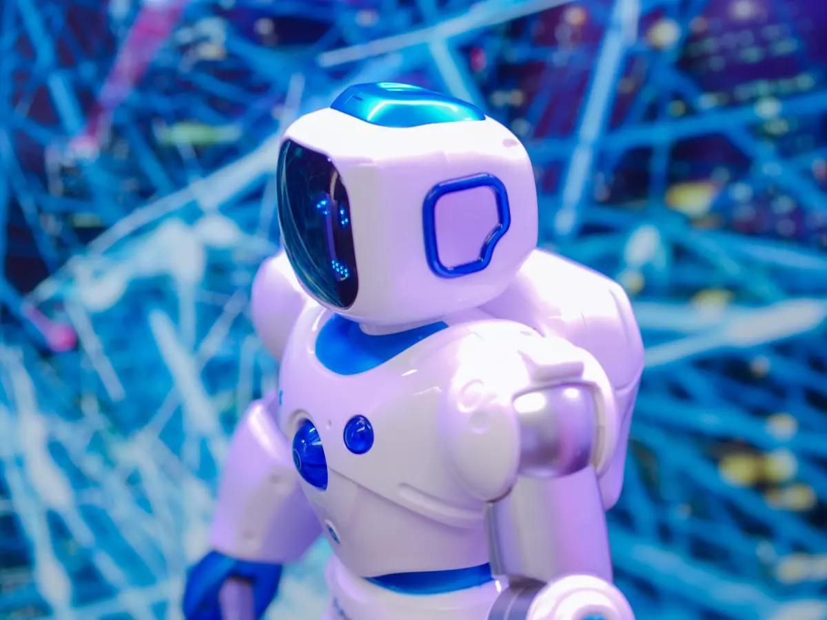 A white humanoid robot with blue accents stands in front of a blue and white abstract background. The robot's head is turned slightly to the left, and its arms are bent at the elbows. The robot's torso is clearly visible, showcasing its design and functionality.