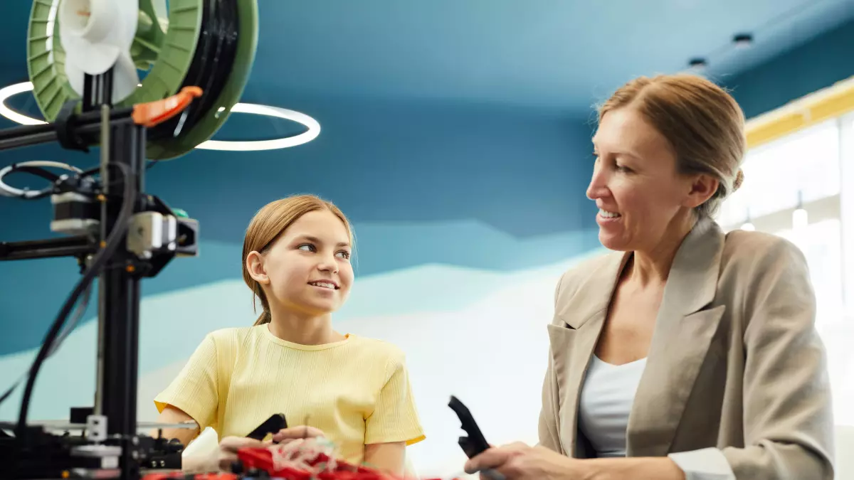 A young girl and an older woman are sitting at a table in front of a 3D printer. The girl is holding a small red object in her hand, and the woman is looking at her with a smile. The 3D printer is in the background, and a light is shining on it.