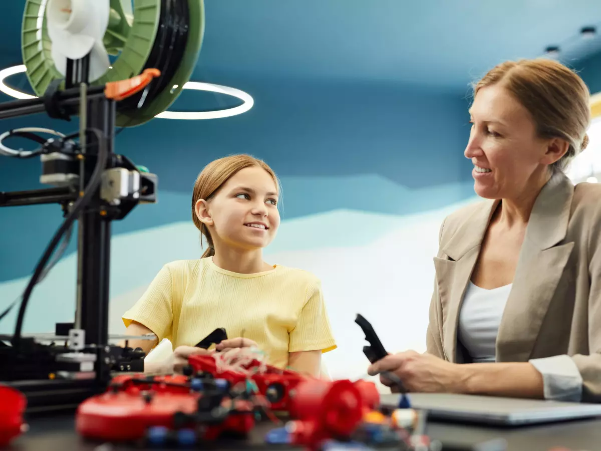 A young girl and an older woman are sitting at a table in front of a 3D printer. The girl is holding a small red object in her hand, and the woman is looking at her with a smile. The 3D printer is in the background, and a light is shining on it.