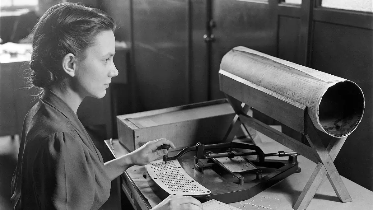 A woman in a shirt is seated in front of a punch card machine, her hands are working on it.