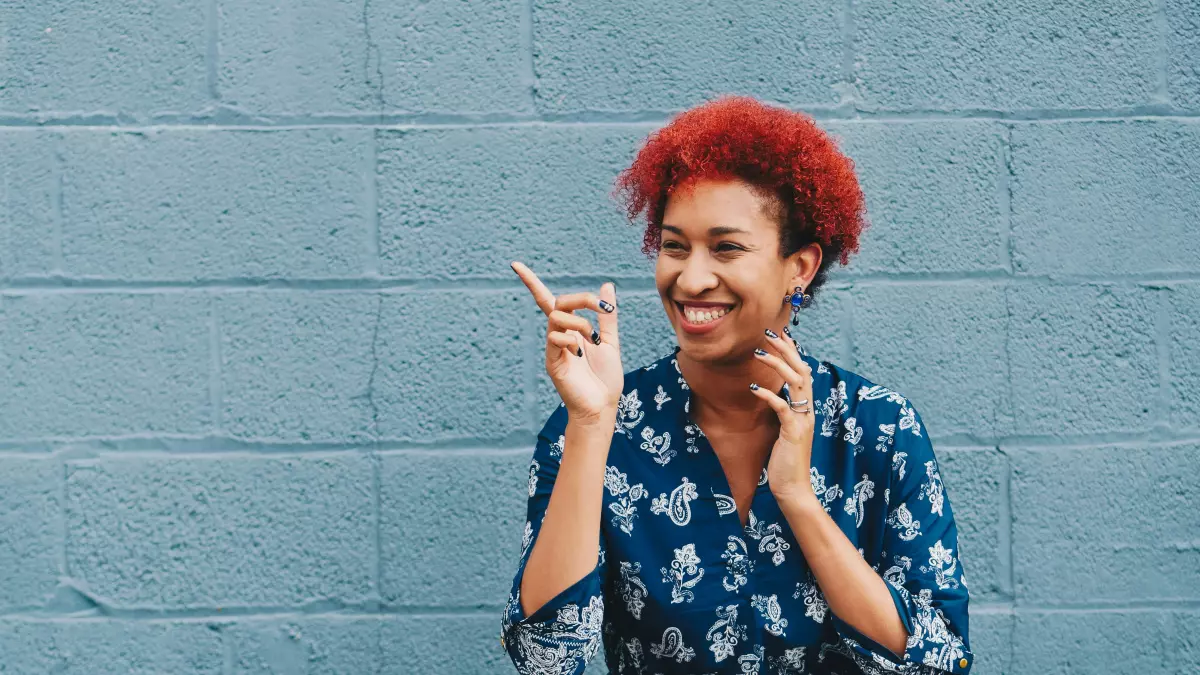 A woman with bright red hair is smiling and pointing to something off-camera.