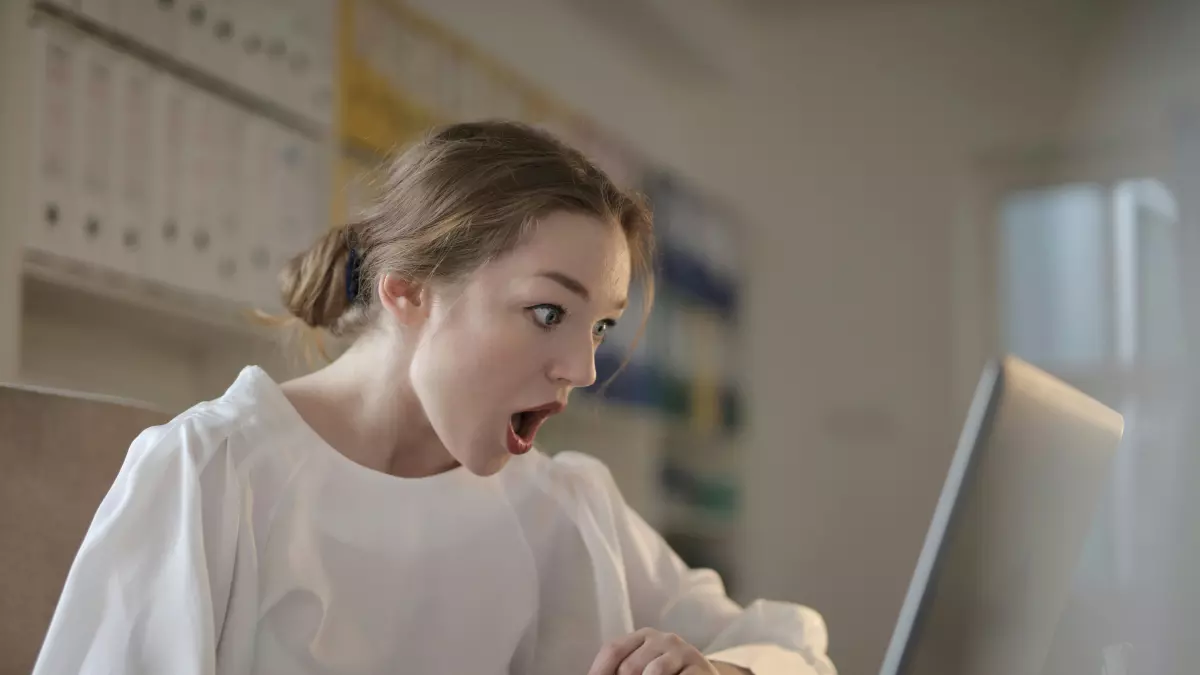 A woman is sitting at a desk and looking at a laptop screen. She has a surprised expression on her face. She is wearing a white shirt and has her hair pulled back.