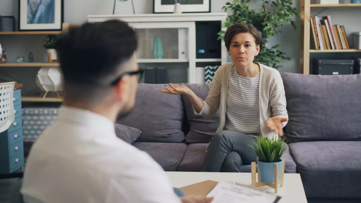 A man and a woman sitting on a couch, having a serious conversation. They are both looking at each other with expressions of interest.