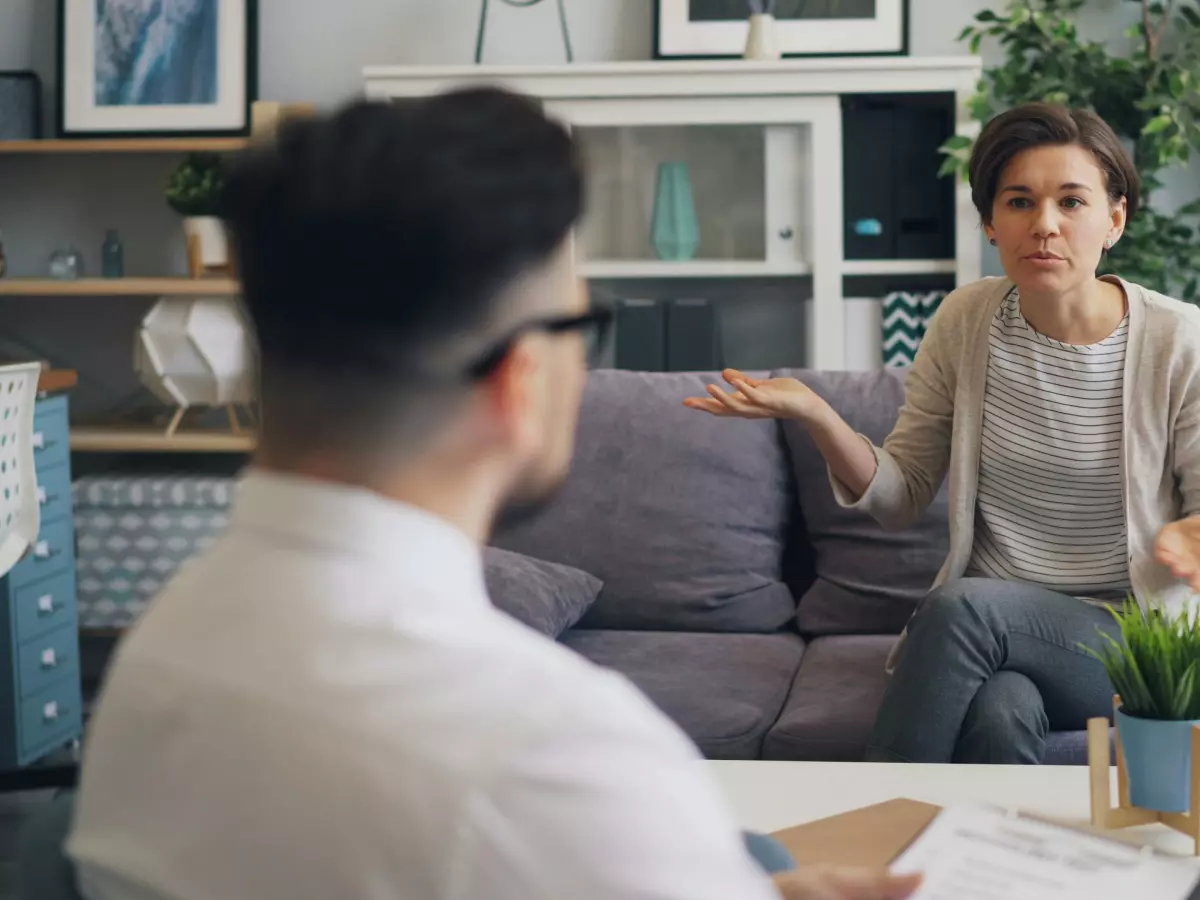 A man and a woman sitting on a couch, having a serious conversation. They are both looking at each other with expressions of interest.