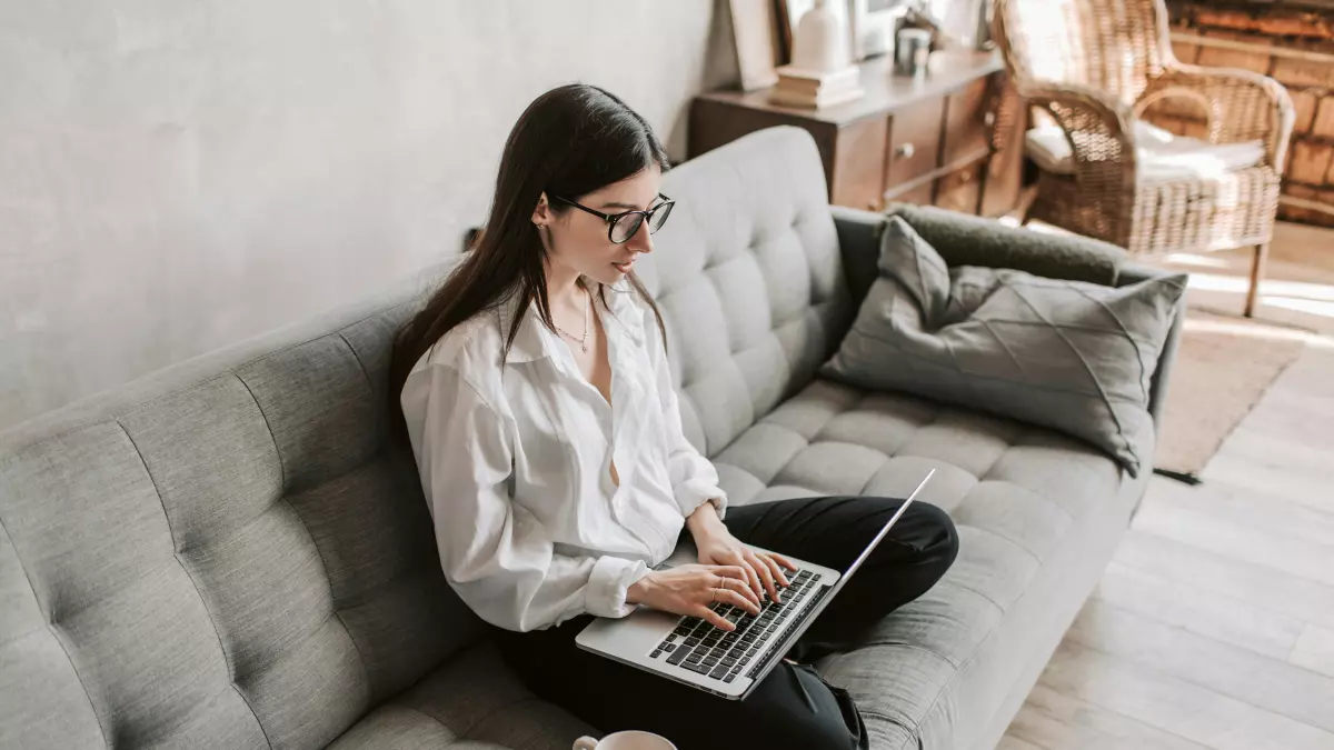 A woman sitting on a couch in a living room, working on a laptop.