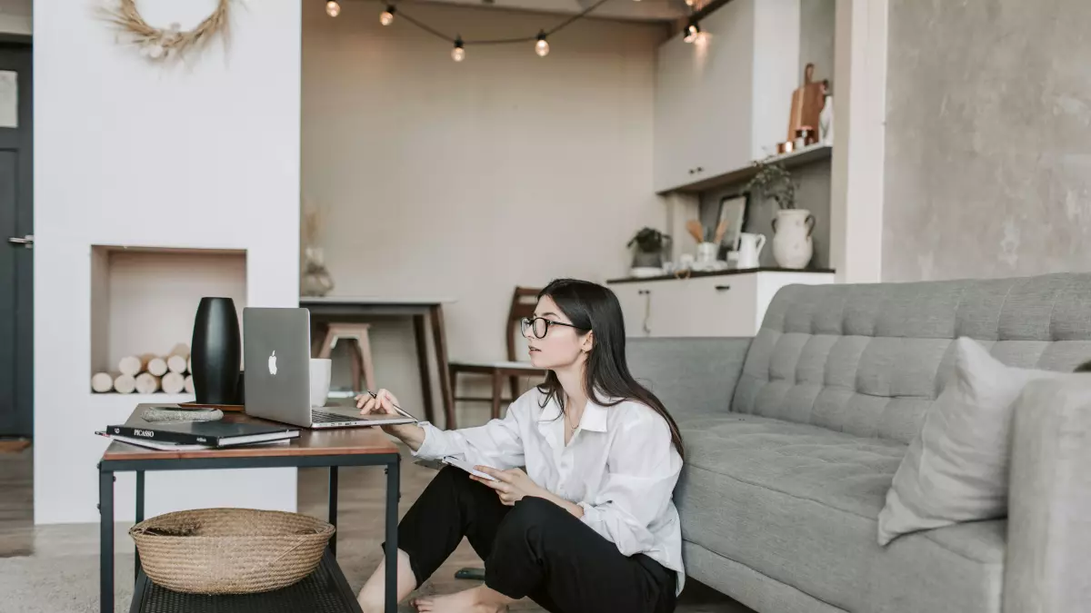 A young woman is sitting on the floor in a living room, working on a laptop. She is wearing a white shirt and black pants. There is a sofa, a coffee table, and a lamp in the background.