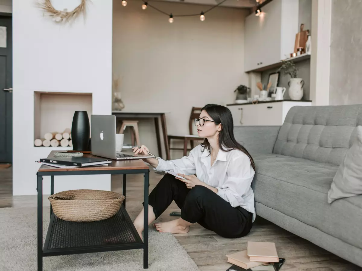 A young woman is sitting on the floor in a living room, working on a laptop. She is wearing a white shirt and black pants. There is a sofa, a coffee table, and a lamp in the background.